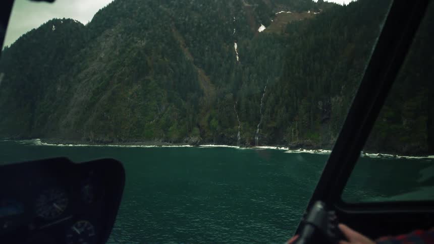 A man flies a helicopter past tree covered coastal hills with waterfalls