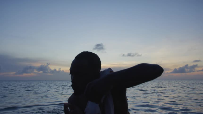 Silhouette of a young shirtless man standing in front of the ocean dancing at sunset