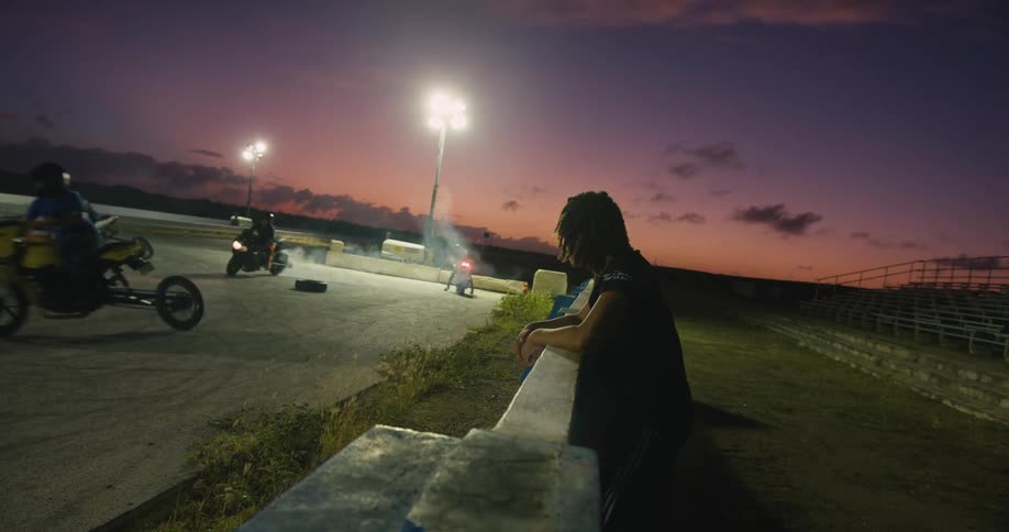 A young man watches a group of motorcyclists ride around a racing strip at a race track at sunset