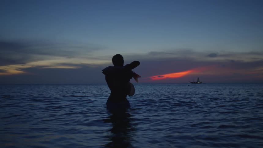 Silhouette of a man standing in the water doing tai chi movements as the sun sets on the horizon