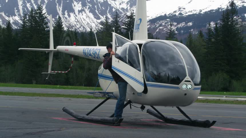 A male pilot climbing into the cockpit of a helicopter parked near a snowy mountain range