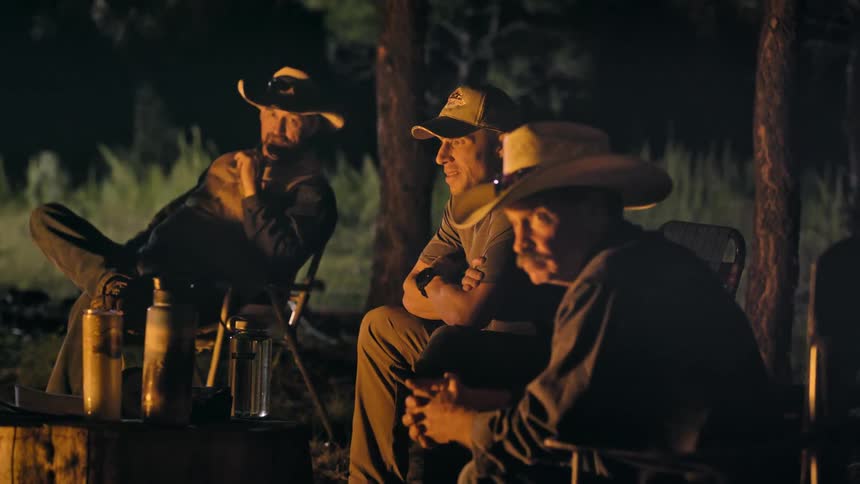 A group of men watch something out of frame while sitting around a campfire at night