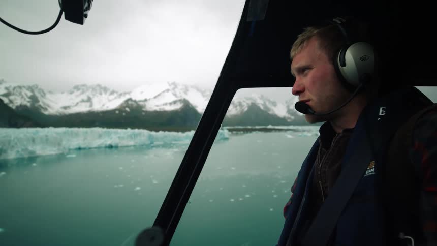 Male pilot with gray headphones and black coat flies helicopter over sea with glaciers next to snowy mountains