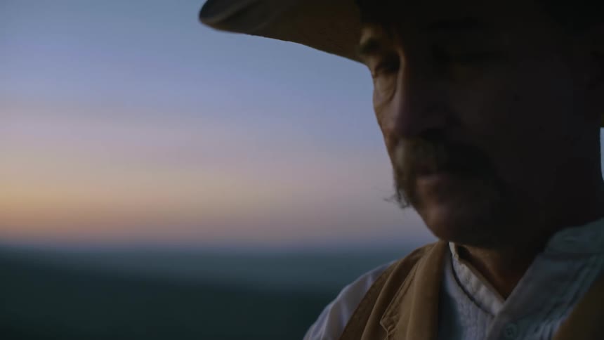 A man in a cowboy hat stands outdoors at sunset
