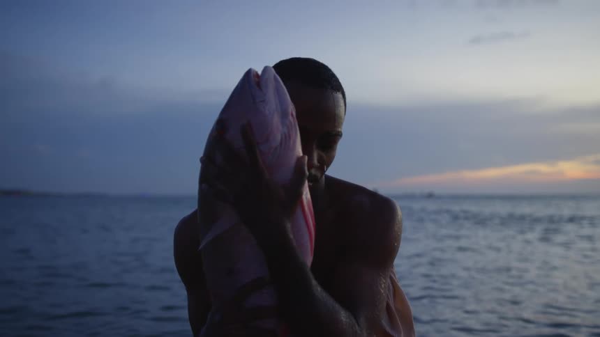 A man standing in the ocean while hugging a large fish to his body opens his eyes to look into the camera
