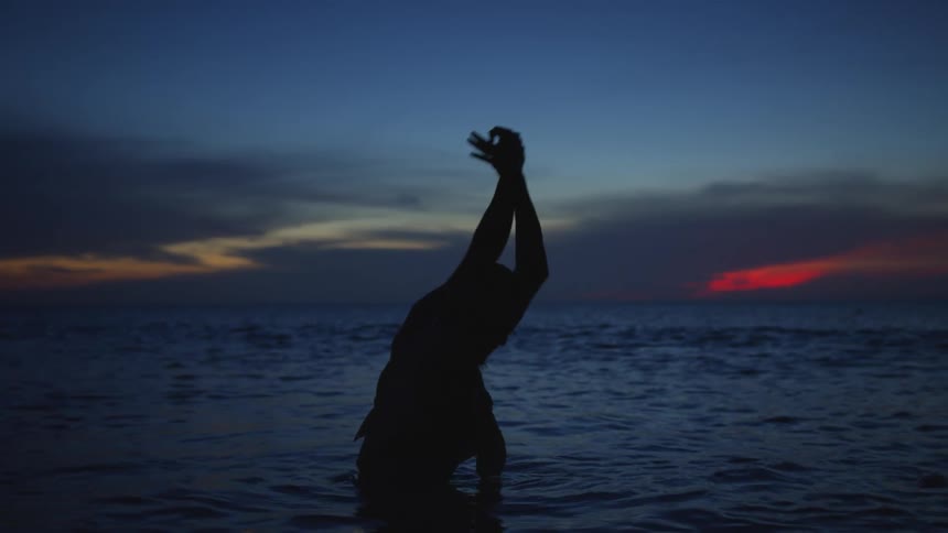 Man standing in ocean raises hands above his head silhouetted against sky and crimson on the horizon