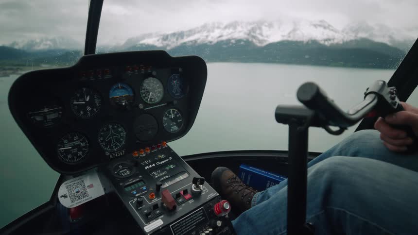Pilot steers helicopter over water with snow covered mountains on shore