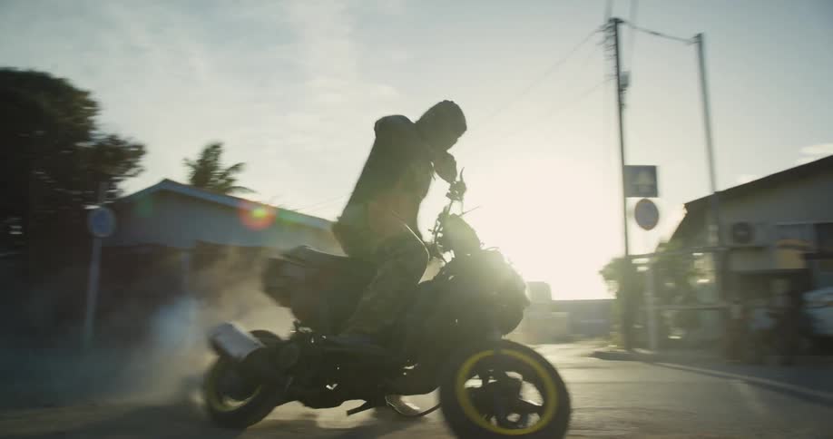 Man wearing mask turns motorcycle in tight spin kicking up dust on road near buildings