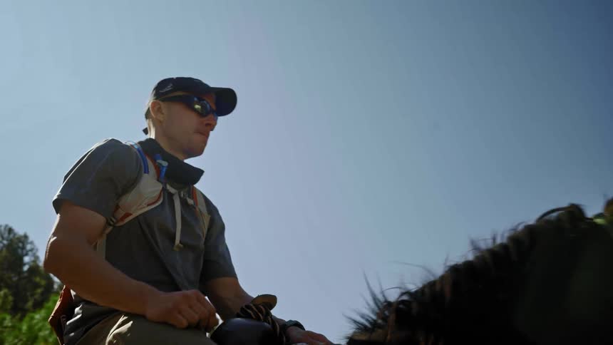 Man with cap sunglasses t shirt and pants rides dark horse under a cloudless sky