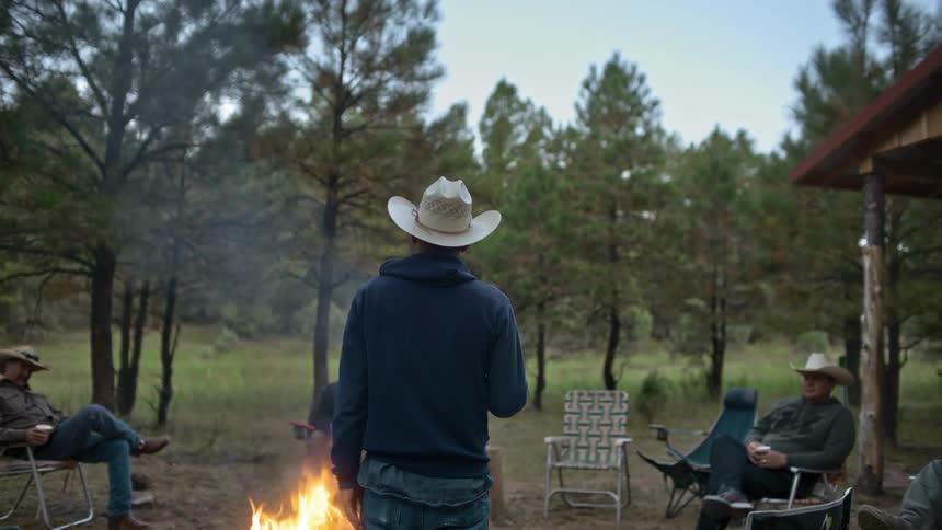 Three cowboys sit in chairs around a fire listening to a cowboy standing and talking