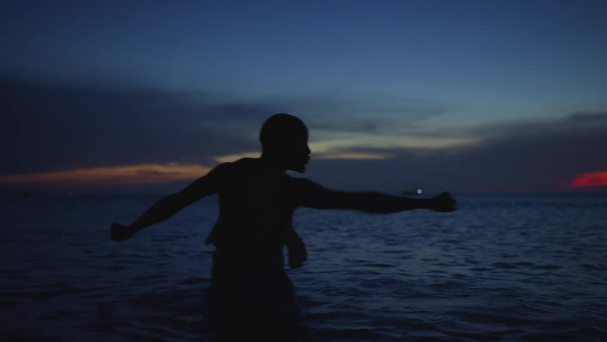 Silhouette of a man standing in the water doing tai chi movements as the sun sets on the horizon