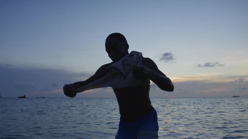 Man stretch top worn around his neck as he stands in ocean with boat in background near horizon