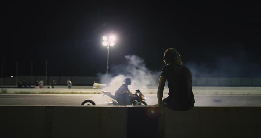 Spectators watch motorcycle racers burn rubber on race track at night