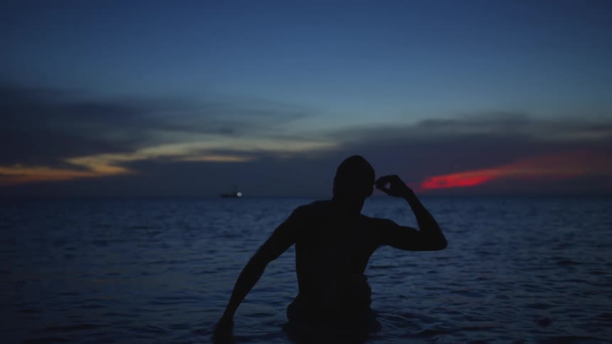 Shirtless man dancing on the beach is silhouetted against the sunset at dawn