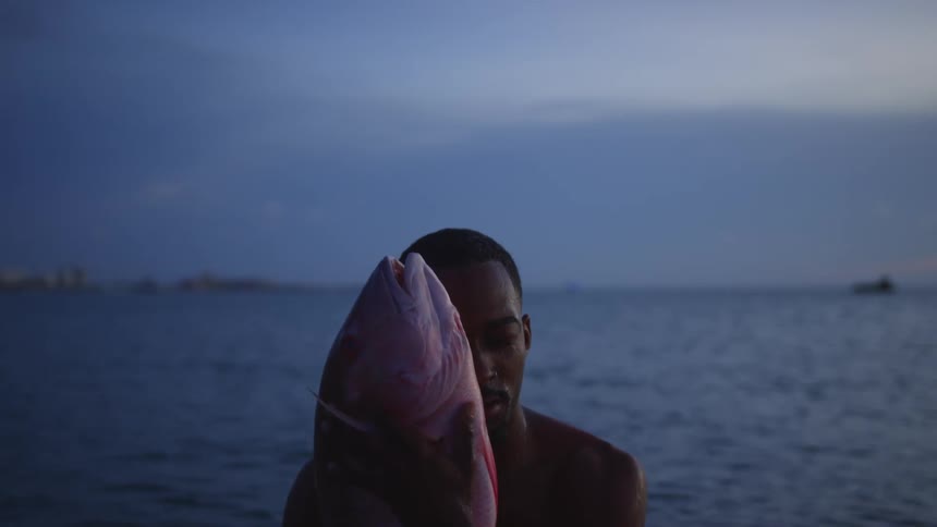 Young shirtless man holding a dead fish standing in front of the ocean at sunset