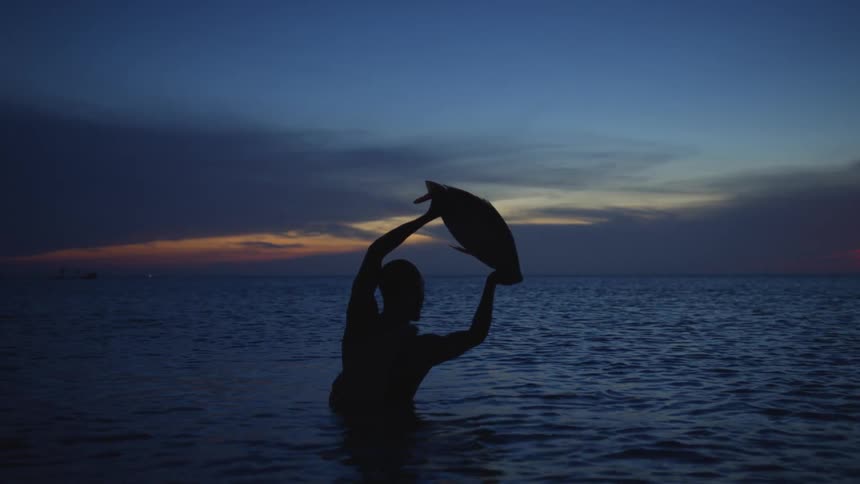 Silhouette of a man standing in the water holding up a large fish in his hands as the sun sets on the horizon