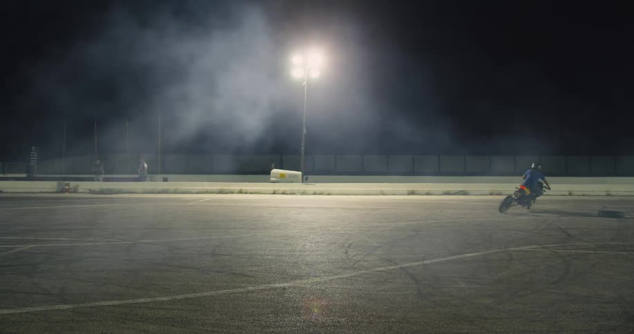 Men drive motorbikes around an arena at night as two other men watch