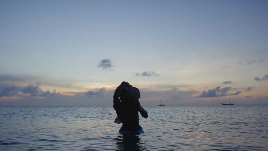 A man standing in the ocean stretches his arms out pulling his shirt and wrapping it around his neck