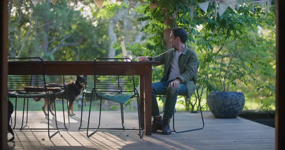A man sitting on his deck pets his dog and throws him a ball before taking a sip from his drink