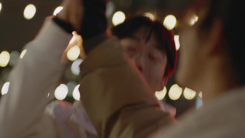 Two friends are having a lively conversation while sitting in a downtown area with glowing string lights and storefronts at night