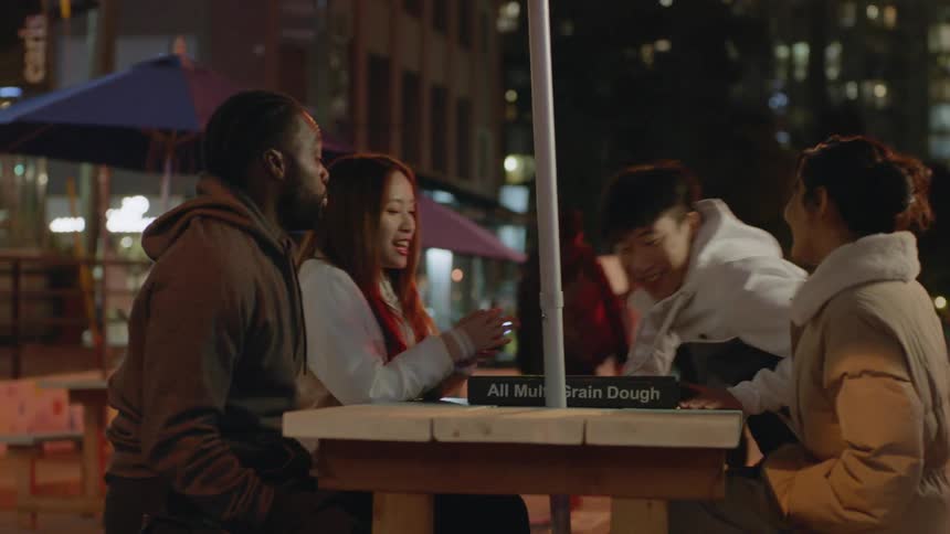 A young man brings a pizza to an outdoor table and a woman claps and he sits down with his three friends