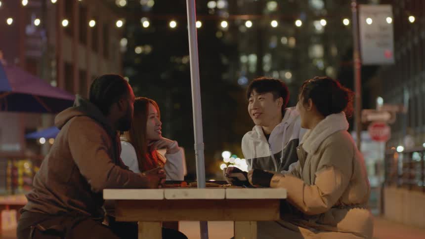 A group of friends talk with one another while sitting at an outdoor table in a city at night