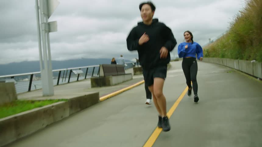 Three people are jogging along a paved pathway near a lake with mountains in the background on an overcast day