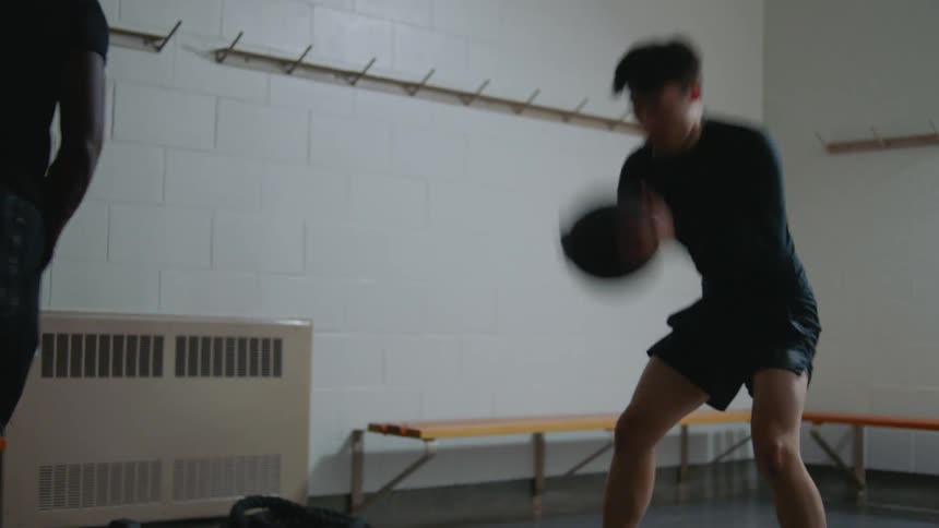 A man demonstrates an exercise ball exercise to a younger man who then performs the exercise