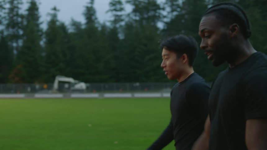 A pair of men talk while walking together across a sports field in the evening
