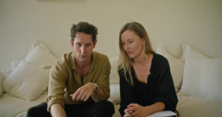 Man with yellow shirt and black pants and blonde woman wearing black blouse sit on beige couch work on laptop in living room at home