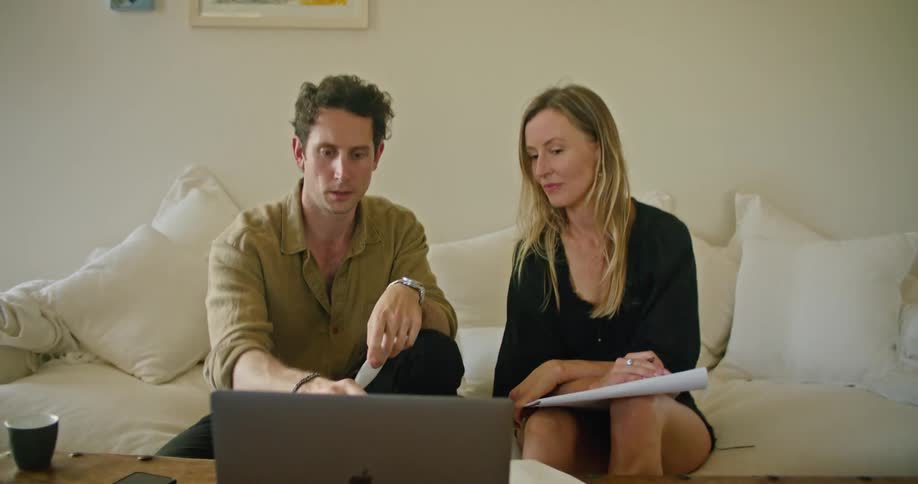 Man and woman sit on couch discussing paper and contents of laptop