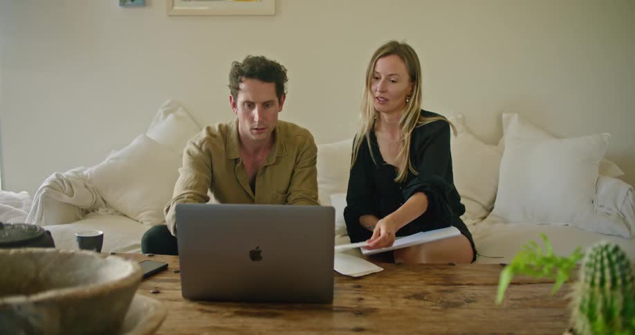 A man and woman review papers while sitting together on a sofa indoors during the day