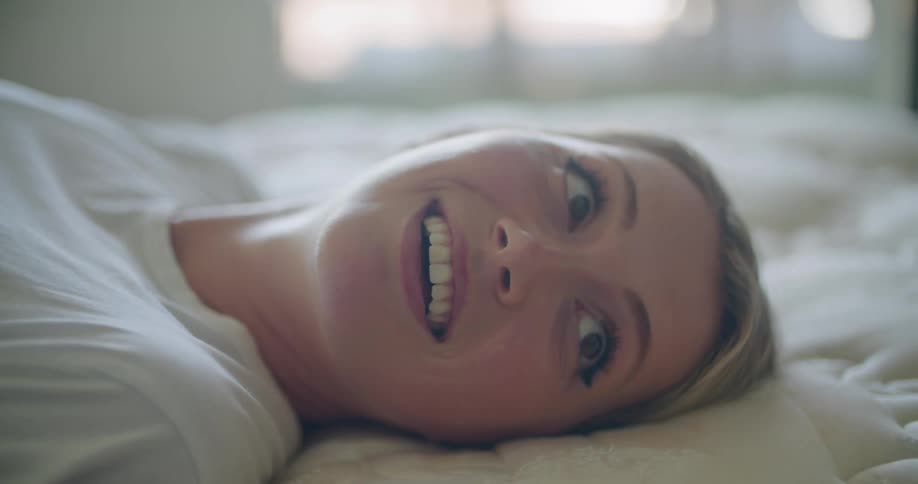 Blonde woman with white t shirt lies on bed laughing at person in bedroom