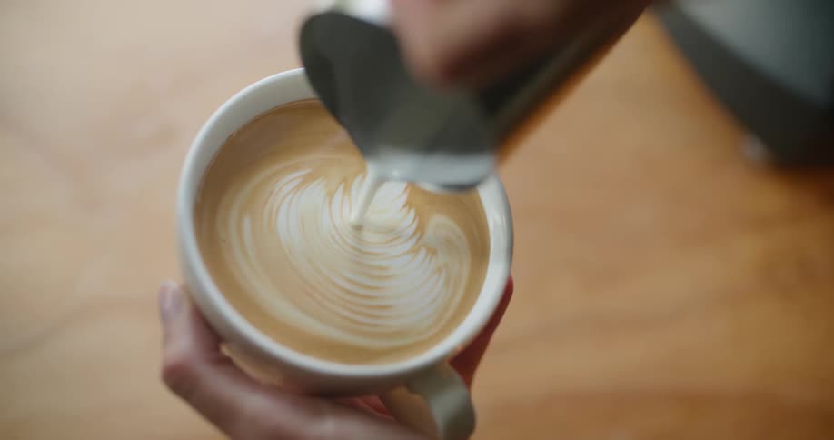 A woman preparing a latte in a white ceramic mug and placing it on a wood table