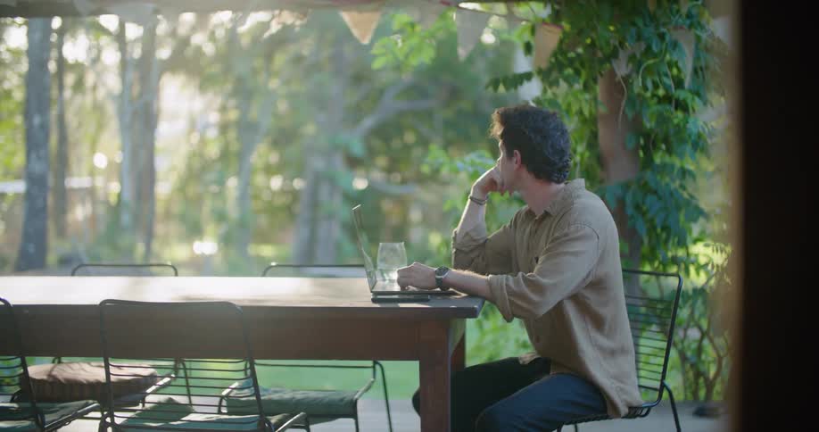 A man sitting at a patio table trying to work on his laptop is distracted by the wind blowing the trees