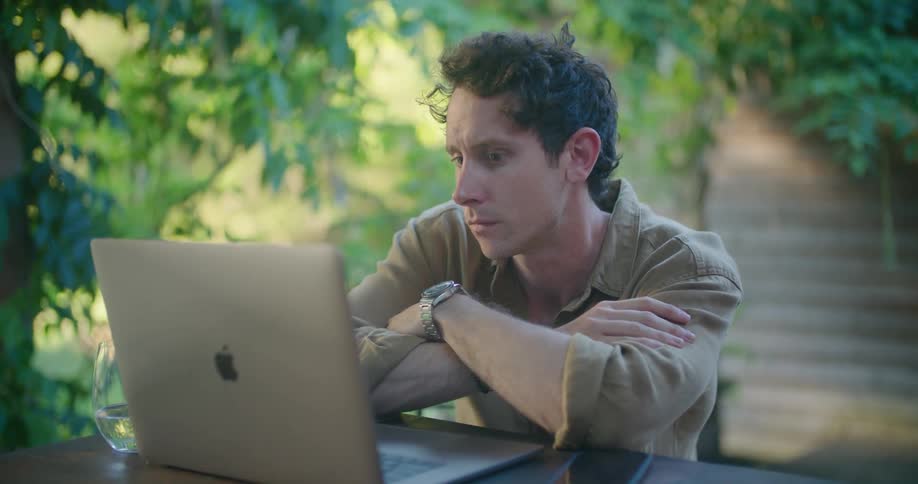 Man with yellow shirt and wristwatch sitting at table thinks before typing on laptop in the green garden