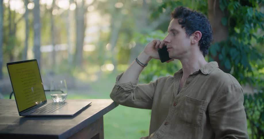 Man sitting at a table on the porch talking on the phone while using a laptop in the morning