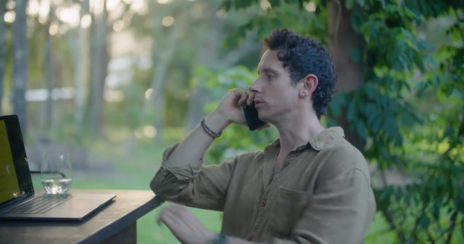 Man sitting at a table in the porch talking on the phone while working on a laptop at morning