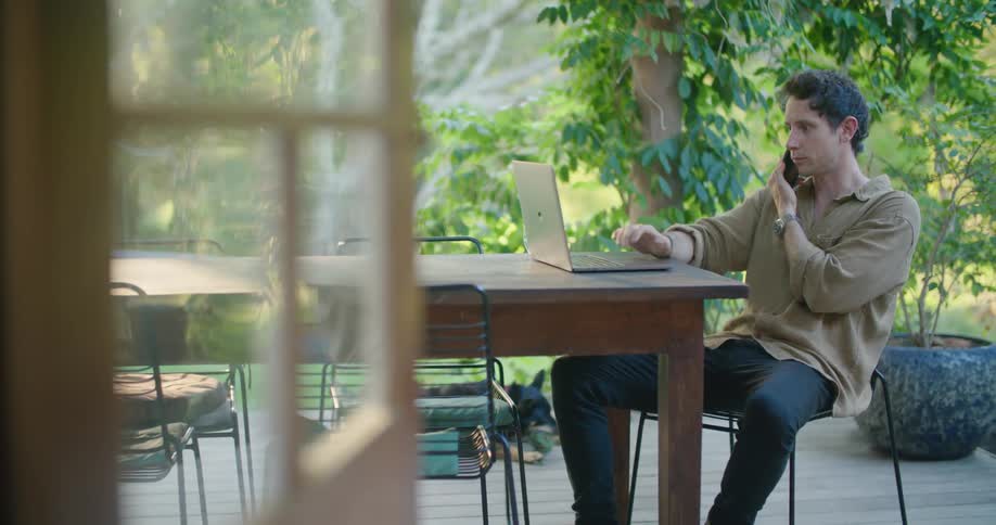 A man speaks on his smartphone while using his laptop at a table outdoors during the day