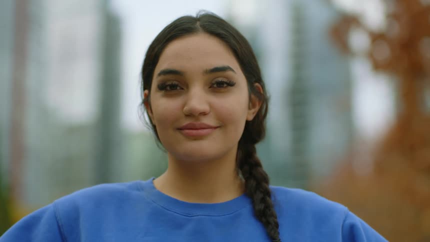 Woman with braids and blue sweater looks at camera with eye contact in public park with high rise buildings in the background