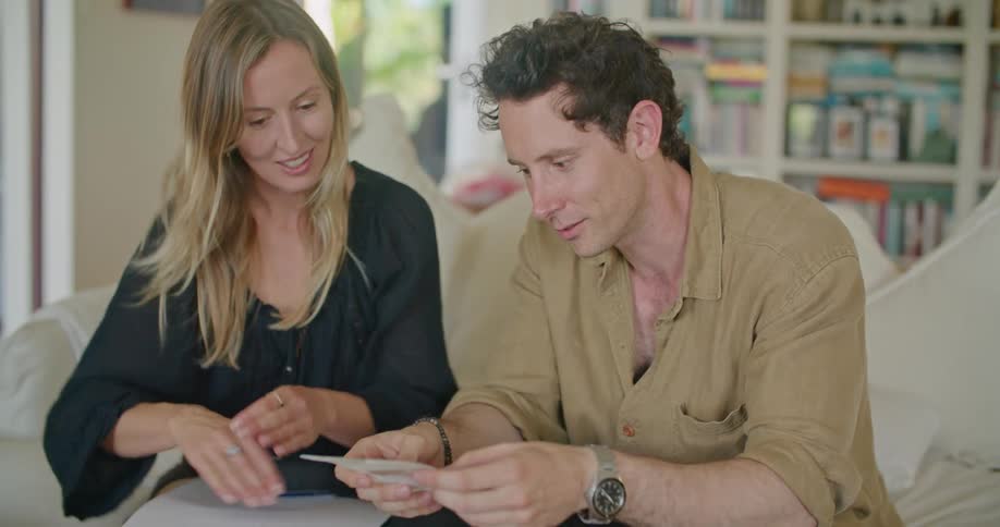 Man and woman look over and discuss flyers stacked on coffee table near laptop