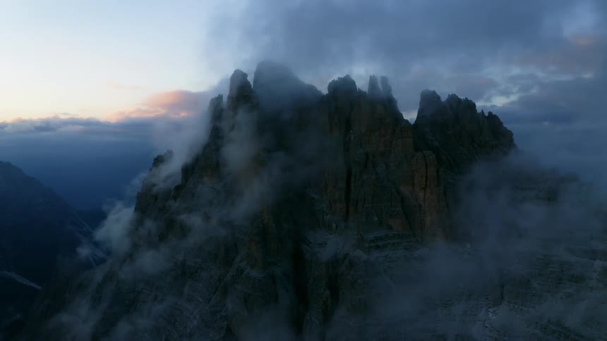 Aerial view of whispy white clouds over the jagged peak of a mountain in the evening