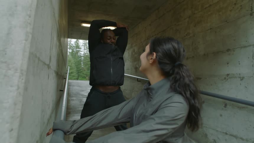 A man stretches with his arms above his head as a woman uses a railing to stretch her legs
