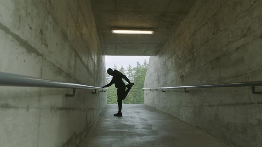 Man hold onto rail in corridor and stretches his leg with conifer trees in background