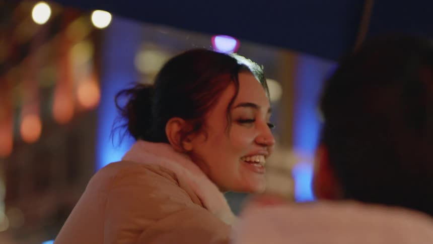Woman wearing collared coat greets friends near bright building at night