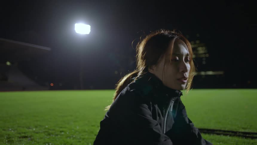 A woman squatting on a sports field at night rests her eyes for a moment before a man offers a hand to help her to her feet