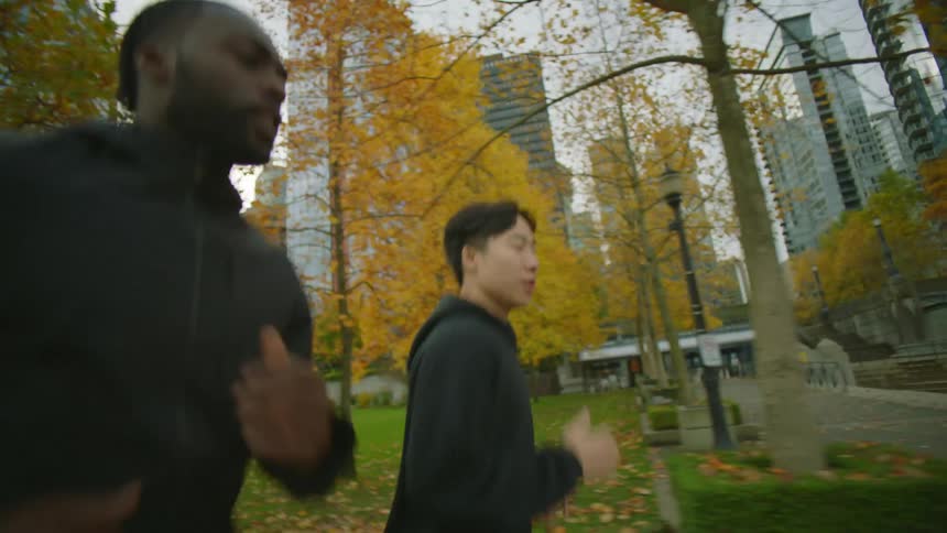 Two men are jogging through a public park in a downtown area of a large city on an overcast day in the fall season