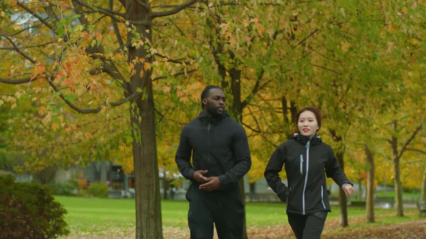 A man and woman are jogging through a park with trees dropping leaves in fall colors