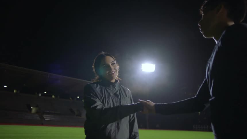 Man and woman catching breath standing on sports field at night clasp hands