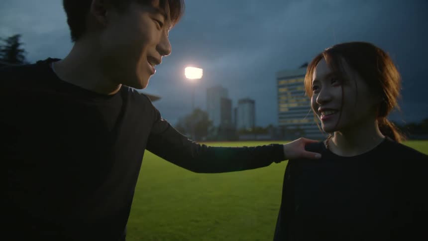 Man talks to woman then hugs her while they are standing on sports field at night